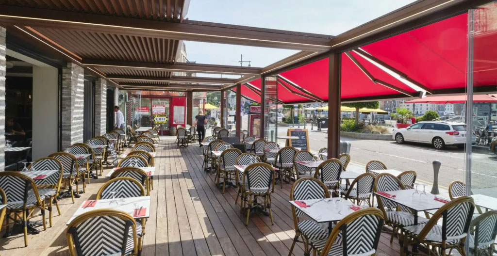 Terrasse semi-fermée de restaurant avec pergola vitrée, tables en U pour événement professionnel, participants en business casual échangeant, décor contemporain épuré, lumière naturelle douce